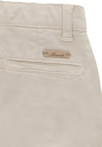 Pantalones beige con una textura suave, que cuentan con un bolsillo trasero y una pequeña etiqueta de cuero etiquetada como "Ananda". Las costuras son sutilmente visibles.