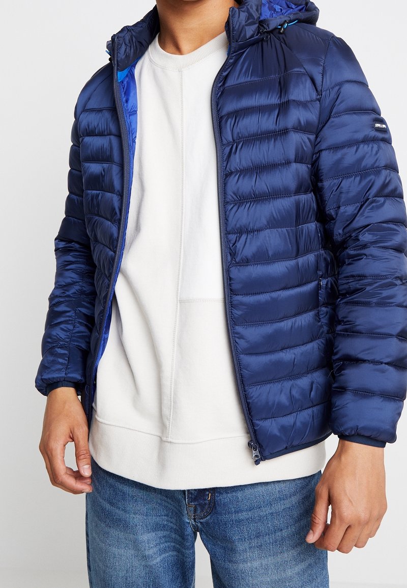 Donkerblauw donsjack met een doorgestikt design, voorzien van een capuchon en een contrasterende voering aan de binnenkant. Ritssluiting aan de voorkant en elastische manchetten.