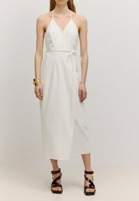 Weißes, halterförmiges Wickelkleid mit tailliertem Gürtel. Hergestellt aus leichtem Stoff, hat es einen tiefen V-Ausschnitt und eine fließende Silhouette. Braune Riemchensandalen.