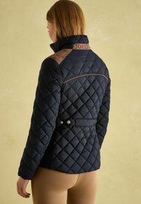 Veste matelassée bleu marine avec col montant, accents en daim sur les épaules et ceinture à la taille ornée de matériel doré. Vue de dos montrant un motif en losanges.