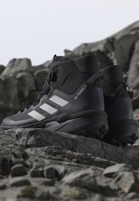 Sorte Adidas Terrex højtop-vandresko med hvide striber, placeret på klippefyldt terræn med sløret klippebaggrund.