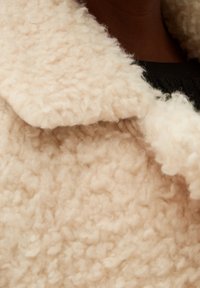 Manteau en polaire couleur crème avec une surface douce et texturée et un large col. Le tissu semble moelleux et confortable, idéal pour se réchauffer.