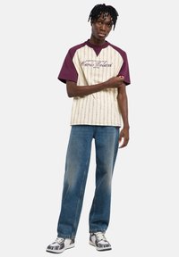 Μπλούζα με κοντά μανίκια σε κρεμ και μπορντό χρώμα με κάθετο ριγέ σχέδιο, κεντημένο κείμενο, συνδυασμένη με ανοιχτό μπλε χαλαρό τζιν και λευκά αθλητικά παπούτσια με σχέδια.