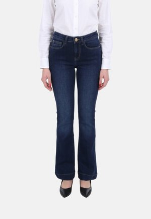 Donna con jeans bootcut blu scuro, camicia bianca abbottonata e tacchi neri a punta, in piedi davanti a uno sfondo bianco.
