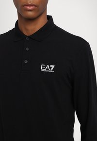 Črna dolga majica s polo ovratnikom iz bombaža. Ima klasičen ovratnik in tri gumbe, ter belo EA7 Emporio Armani logo na prsih.