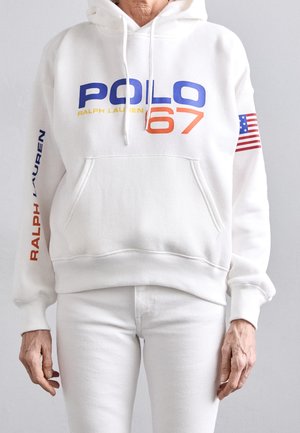 Osoba nosząca białą bluzę z kapturem z napisem „POLO 67” w kolorach niebieskim i pomarańczowym, tekstem „RALPH LAUREN” na rękawie oraz naszywką z flagą USA na prawej ręce.