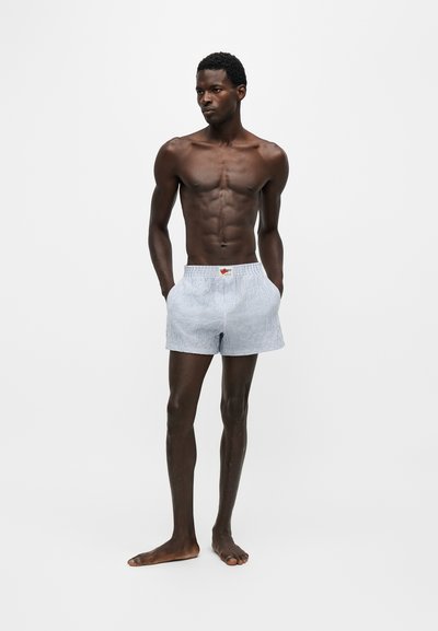 Homme mince à la peau foncée, debout pieds nus, portant un short rayé bleu clair, les mains dans les poches, sur un fond blanc uni.