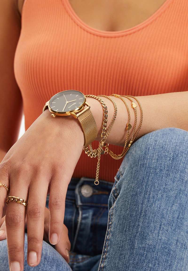 Goldene Mesh-Armbanduhr mit schwarzem Ziffernblatt, übereinander getragene goldene Kettenarmbänder und Ringe auf brauner Haut. Orangefarbener gerippter Oberteil und Jeans sichtbar.