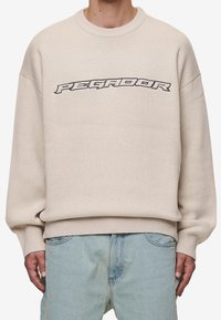 Beige stickad sweatshirt med rund halsringning, sänkta axlar och strukturerad finish. Framsidan har designen "PEGADOR" i svart.