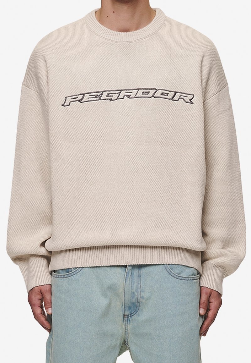Beige stickad sweatshirt med rund halsringning, sänkta axlar och strukturerad finish. Framsidan har designen "PEGADOR" i svart.