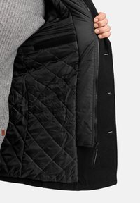 Schwarzer Mantel mit einem gesteppten schwarzen Innenfutter. Verfügt über einen Reißverschluss und eine kleine Tasche, strukturierten Wollaußenbereich und subtile Nahtdetails.