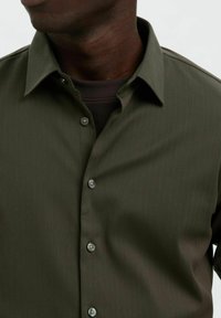 Ciemnooliwkowa koszula zapinana na guziki z subtelny wzorem pionowych pasków, z klasycznym kołnierzykiem i szarymi guzikami, noszona na ciemnym topie.