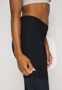 Svarta träningsleggings med hög midja, sidoficka med dragkedja och strukturerade paneler på underbenen. Tillverkade av stretchigt, fukttransporterande material.