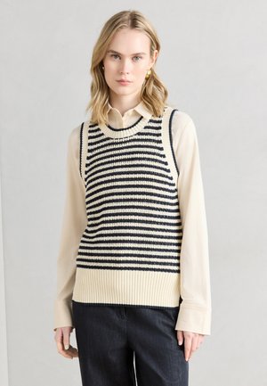 Mladá žena nosiaca zlaté kruhové náušnice, krémovú košeľu na gombíky a krémový a čierny pruhovaný pletený bezrukávový sveter, stojaca na sivom pozadí.