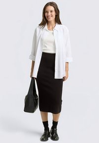 Biała koszula na guziki, czarna dopasowana spódnica, czarne skarpetki oraz czarne buty. Modelka trzyma czarną torbę typu tote, stoi na neutralnym tle.