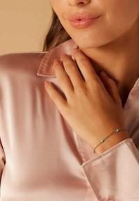 Gouden kettingarmband met kleine turquoise kralen. Draagt een hand die een roze satijnen shirt met kraag aanraakt, wat de gladde textuur laat zien.
