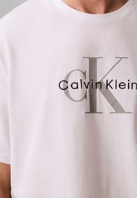 Hvit bomulls-T-skjorte med "CalvinKlein"-logo i grått, med fremtredende "CK"-design. Klassisk rund hals og korte ermer.
