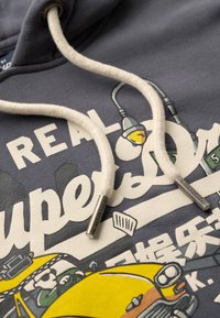 Felpa grigia scura con stampa grafica e testo bianco. Presenta cordini beige con punte in metallo etichettate "SUPERDRY". Texture morbida.
