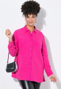 Helles Pink, knopfbares Hemd aus strukturiertem Stoff, mit zwei Brusttaschen und einem etwas längeren Hinterteil. Kombiniert mit einer kleinen metallischen Handtasche.