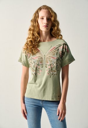 Mujer joven con cabello rizado que lleva una camiseta verde con un gran diseño de mariposa y jeans azules, de pie frente a un fondo liso.