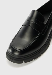 Mocassino in pelle nera con una texture liscia, caratterizzato da una fascia decorativa sulla parte superiore e una suola in gomma spessa con un motivo battistrada.