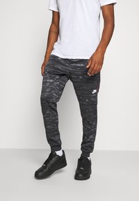 Černé tepláky s texturovaným grafickým designem, elastickými manžetami, bočními pruhy a logem Nike. Kombinovány s černými teniskami a bílým tričkem.