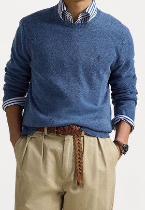 Man draagt een blauwe trui met ronde hals over een blauw-wit gestreept overhemd, beige geplooide broek, bruine gevlochten riem en een polshorloge.