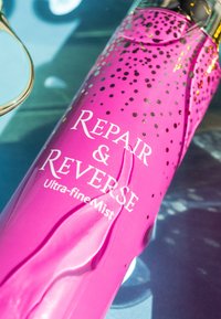Lyserød flaske med guldprikker og prægede bogstaver, mærket "Repair & Reverse Ultra-fine Mist." Glat tekstur, slank cylinderform.