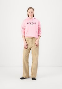 Felpa con cappuccio rosa con "NEW YORK" in nero sul davanti, abbinata a pantaloni larghi beige chiaro e scarpe nere.