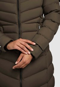 Roku rokas pielāgo tumši olīvu krāsas siltā aksesuāra zipu pie piedurknes, kuram ir melns priekšējais zip un redzama zīmola birka.