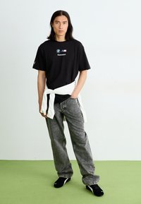 Sort kortærmet t-shirt med BMW Motorsport-logo, parret med grå jeans og sorte sneakers. Hvid sweater bundet om taljen.