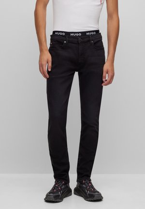 Mand iført sorte slim-fit jeans, sorte sneakers og hvid ærmeløs top med synlig "HUGO" mærket taljebånd.