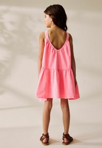 Vestido sin mangas de un brillante color rosa, con una profunda espalda en V, cintura fruncida y falda acampanada. Se lleva con sandalias marrones de tiras.