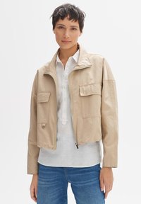 Beige verkürzte Jacke mit zwei frontalen Taschen, Reißverschluss und Stehkragen. Getragen über einem weißen Hemd mit Knöpfen und blauen Jeans.