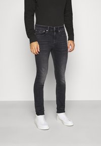 Jeans negros ajustados con detalles desgastados, diseño de cinco bolsillos y acentos metálicos, combinados con zapatillas altas blancas.