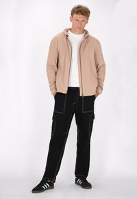 Beige rits hoodie met grijze accenten, gecombineerd met zwarte cargobroeken met witte steken. Zwarte en mintgroene sneakers maken de outfit compleet.