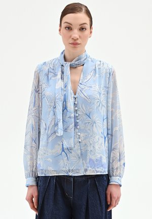Femme portant un chemisier transparent à fleurs bleu clair avec un foulard noué au cou, un devant boutonné et des manches longues, associé à un pantalon sombre plissé.