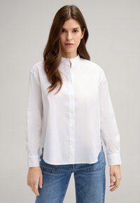 Weißes Langarmhemd mit Stehkragen, mit Knopfverschluss vorne und seitlichen Schlitzen. Hergestellt aus glattem Stoff; kombiniert mit blauen Jeans.
