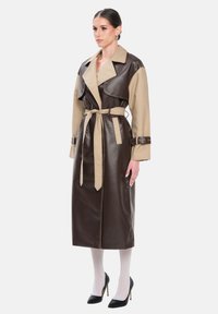 Trench-coat bicolore en similicuir marron et beige avec une ceinture à la taille, un large col et des manches longues. Talons hauts noirs et collants blancs.