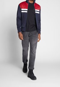 Chaqueta navy con cremallera y hombros rojos, combinada con una camiseta negra, jeans grises y botas negras.