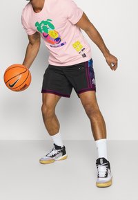 Camiseta rosa con diseño gráfico colorido, pantalones cortos negros con acentos morados, y zapatillas deportivas en blanco y negro, sosteniendo una pelota de baloncesto.