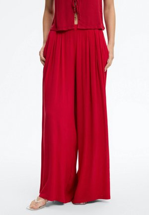 Mujer con pantalones anchos de pierna ancha y pliegues de color rojo brillante, combinados con una camiseta a juego y sandalias con tiras transparentes, de pie frente a un fondo blanco.