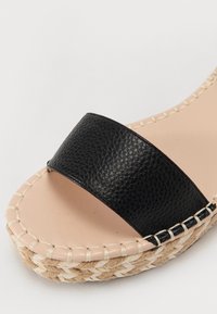 Gros plan d’une sandale espadrille beige avec semelle tissée naturelle et large bride en cuir noir texturé sur la zone des orteils.