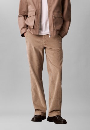 Homme portant un pantalon beige en velours côtelé, une veste marron avec des poches, une chemise blanche et des chaussures en cuir noires, debout les mains dans les poches.