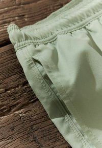 Világos zöld szövet rövidnadrág, rugalmas derékkal, oldalsó varratokkal és sima tapintással, egy fa felületen pihen.