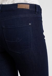 Vaqueros de denim azul oscuro con bolsillo trasero con diseño bordado y una etiqueta de marca de cuero marrón en la cintura.