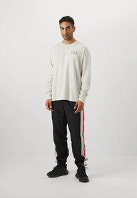 Világosszürke hosszú ujjú ing "Under Armour" logóval, fekete nadrággal, amelyen vörös és fehér oldalsávok vannak, és fekete sportcipővel viselve.