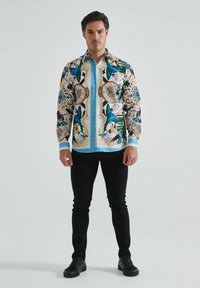 Chemise imprimée colorée avec des motifs bleus, verts et dorés, à manches longues et avec un devant à boutons. Associée à un pantalon noir ajusté et des chaussures.