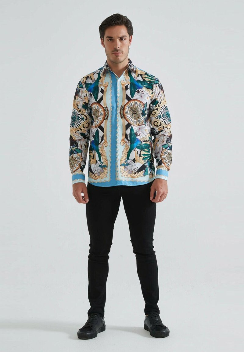 Chemise imprimée colorée avec des motifs bleus, verts et dorés, à manches longues et avec un devant à boutons. Associée à un pantalon noir ajusté et des chaussures.