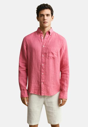 Homme portant une chemise rose à manches longues boutonnée et un short beige clair, debout devant un fond blanc uni.